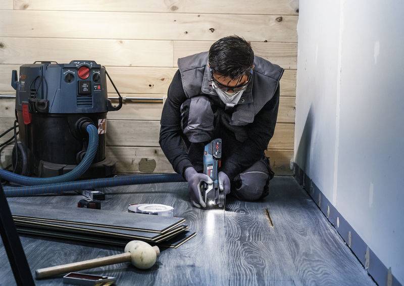 Eine Person mit Schutzbrille und Maske arbeitet kniend mit einem elektrischen Werkzeug an einem Holzboden, ein Staubsauger im Hintergrund.