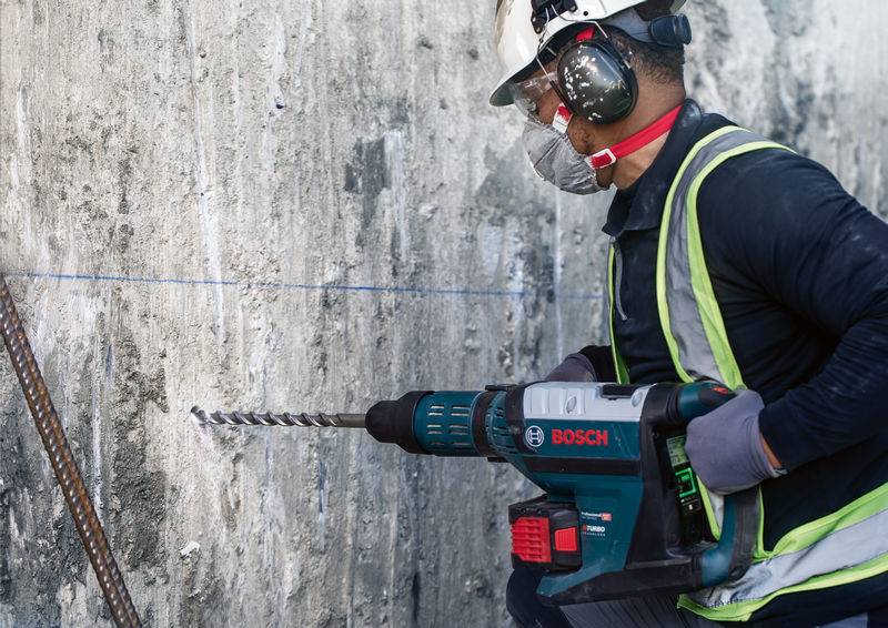 Eine Person mit Schutzkleidung und Gehörschutz bohrt mit einer Bosch-Bohrmaschine in eine Betonwand.