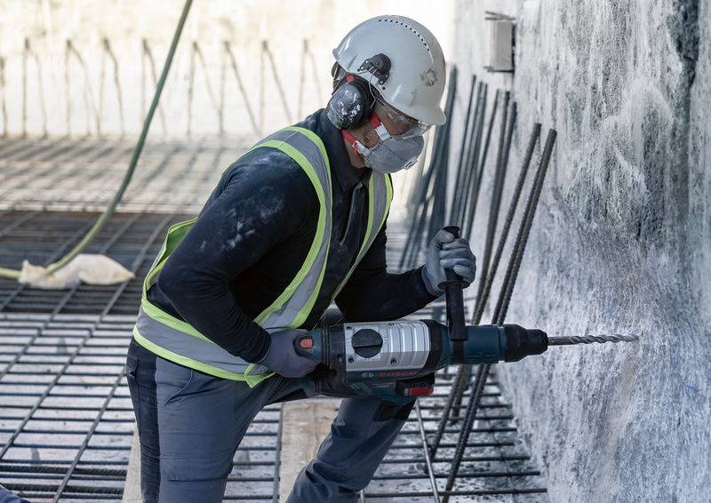 Ein Bauarbeiter mit Schutzhelm, Schutzbrille und Weste bohrt mit einer elektrischen Bohrmaschine in eine Betonwand.