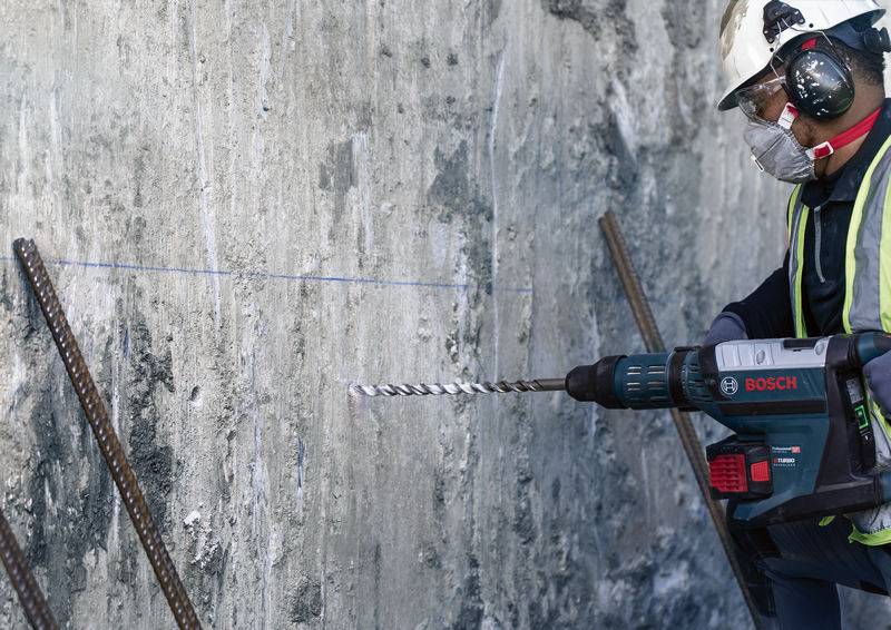 Ein Bauarbeiter mit Schutzkleidung bohrt mit einer Bosch-Bohrmaschine in eine Betonwand. Eisenstangen sind zur Verstärkung sichtbar.