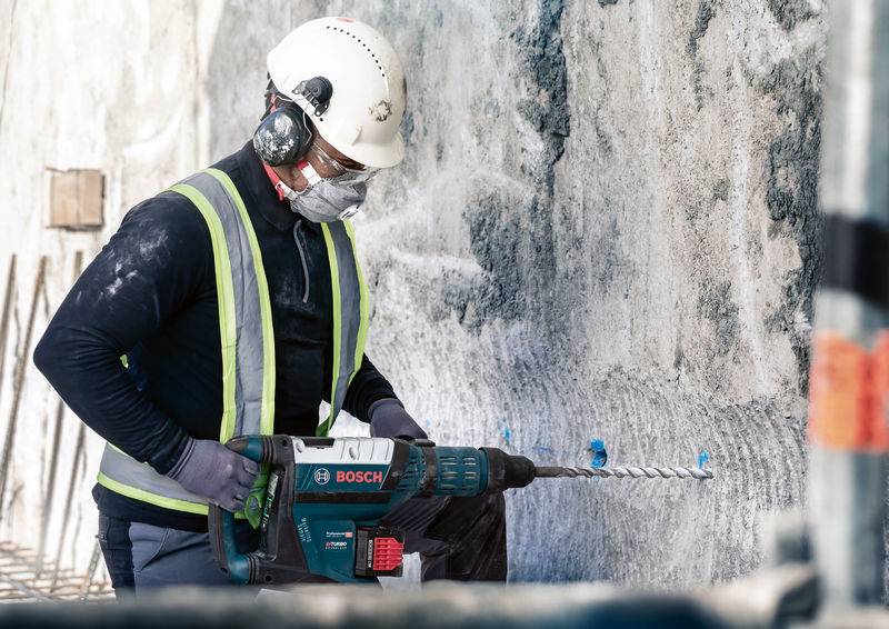 Arbeiter mit Schutzkleidung und Helm benutzt Bohrmaschine, um Betonwand zu durchbohren. Baustellenumgebung, Betonfragmente fliegen.