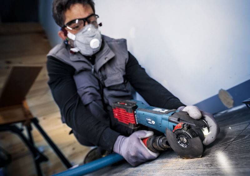 Ein Arbeiter in Schutzkleidung schneidet mit einer elektrischen Schleifmaschine Holz. Er trägt Staubmaske und Schutzbrille.