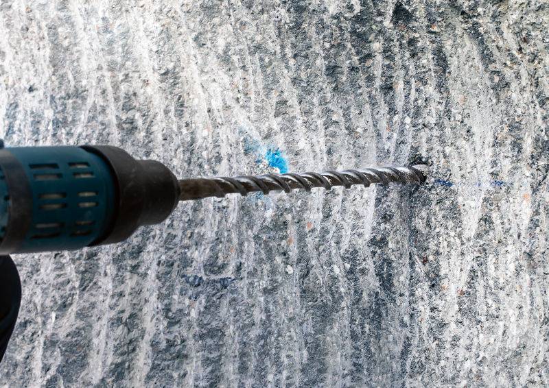 Eine Bohrmaschine bohrt ein Loch in eine graue Betonwand. Die Spitze des Bohrers ist zur Hälfte im Material versenkt.