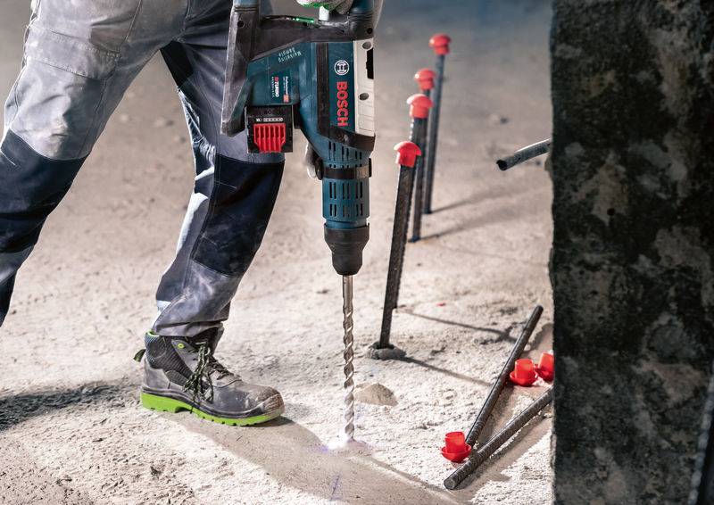 Eine Person bohrt mit einem Bosch-Bohrhammer in einen Betonboden, umgeben von freiliegenden Metallstäben und roten Plastikabdeckungen.