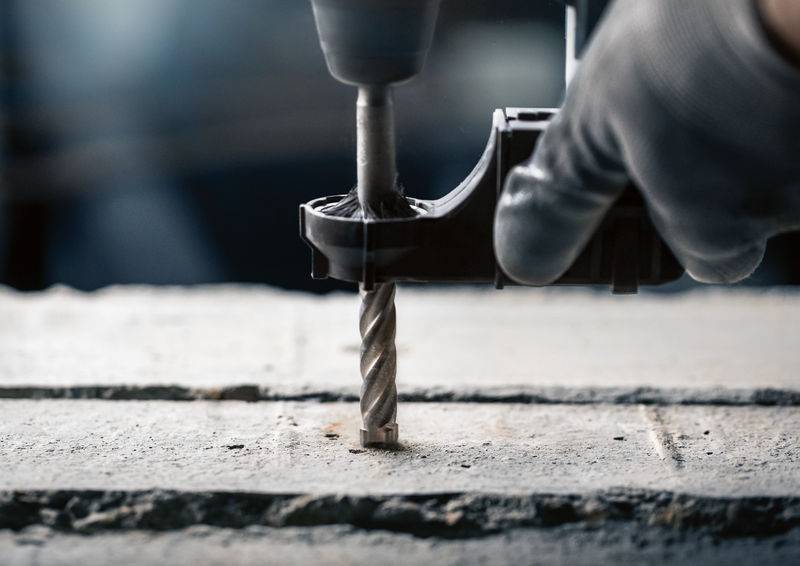 Hand mit Handschuh hält eine Bohrmaschine beim Bohren eines Lochs in eine Betonoberfläche. Staubpartikel sind sichtbar.