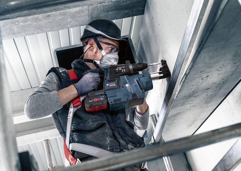 Ein Arbeiter bohrt mit einer Maschine in Beton, trägt Schutzbrille und Helm, befindet sich auf einer Leiter in industrieller Umgebung.