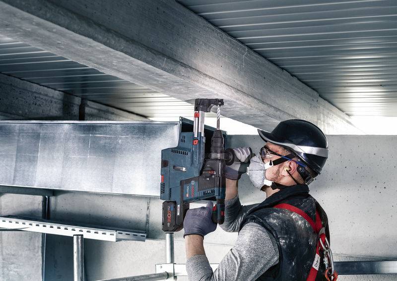 Ein Arbeiter mit Schutzhelm und Maske verwendet ein Elektrowerkzeug, um an einem Luftkanal in einem industriellen Umfeld zu arbeiten.