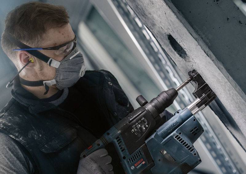 Ein Bauarbeiter trägt Schutzbrille und Maske, benutzt einen Bohrhammer an einer Betonwand. Fokus auf Sicherheitsausrüstung und Präzision.