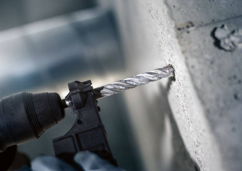 Eine Person bohrt mit einem Bohrer ein Loch in eine Betonwand. Der Bohrer hat ein gedrehtes Metallbohrfutter, das Staub erzeugt.