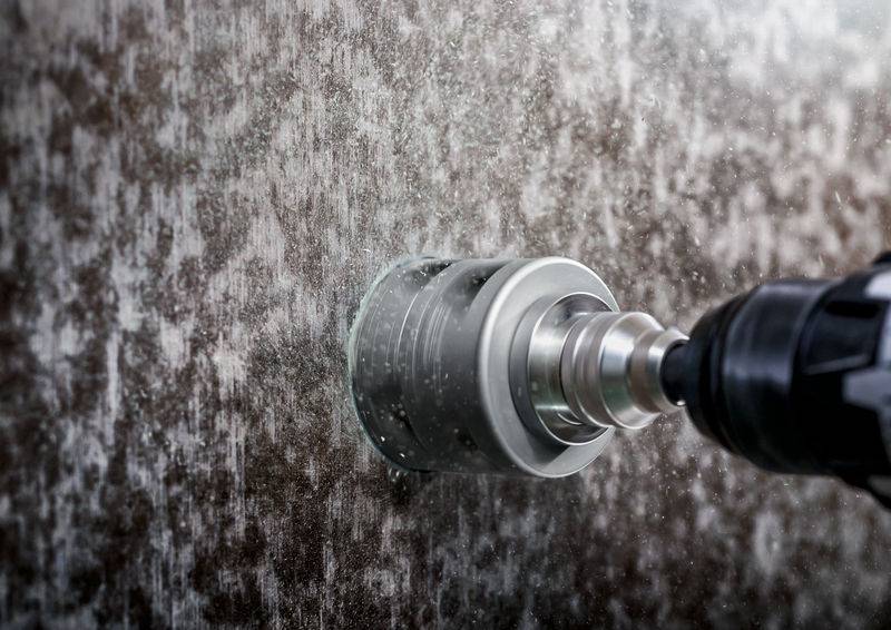 Ein elektrischer Bohrer mit rundem Aufsatz bohrt ein Loch in eine dunkle Steinoberfläche. Wasserspritzer erzeugen einen feuchten Effekt.
