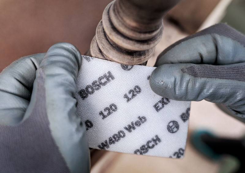 Eine Person trägt graue Handschuhe und schleift ein Holzstück mit einem Bosch Schleifpapier. Das Schleifmittel hat eine Körnung von 120.