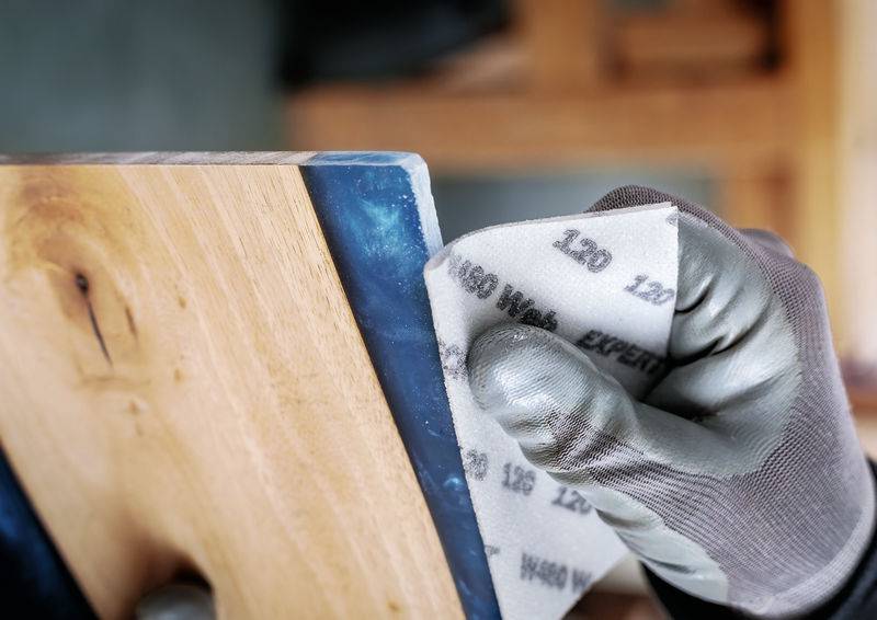 Eine Person schleift mit einem Schleifpapierblock die Kante eines Holzstücks. Sie trägt Handschuhe.