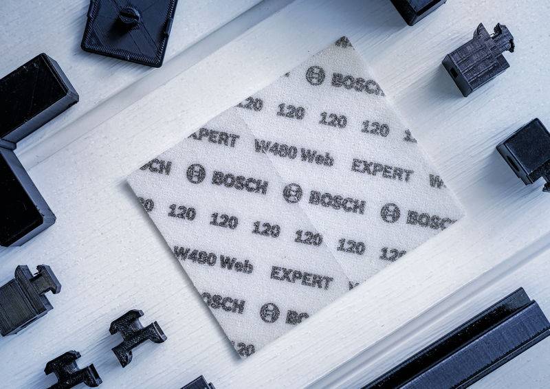 Schleifblatt von Bosch mit der Aufschrift 'EXPERT', '120', und 'W480' liegt auf hellem Hintergrund, umgeben von schwarzen Kunststoffteilen.