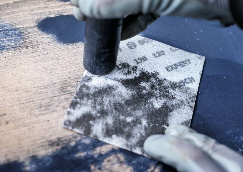 Ein Handschuh hält ein schwarzes Schleifpapier mit der Aufschrift 'EXPERT 150 120', während Holzoberfläche bearbeitet wird.