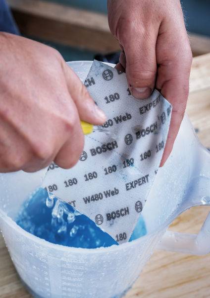 Zwei Hände tauchen ein Schleifpapier der Marke Bosch in einen Krug mit blauer Flüssigkeit.