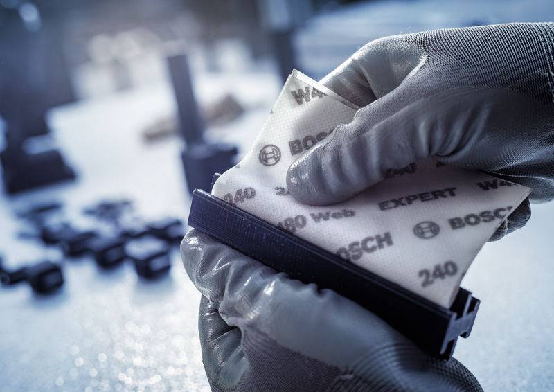 Eine Nahaufnahme von Händen in Arbeitshandschuhen, die ein mit 'Bosch' markiertes Schleifpapier an einem Werkzeug befestigen.