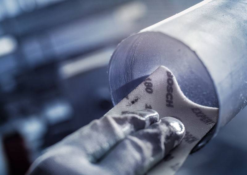 Ein Handschuh hält ein Schleifpapier, das auf eine Metalloberfläche gelegt wird, vermutlich um diese zu glätten oder zu polieren.