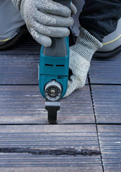 Person benutzt ein elektrisches Werkzeug, um Holzplatten zu schneiden. Nahaufnahme von Händen in Arbeitshandschuhen und dem Werkzeug auf dem Boden.