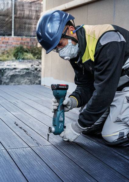 Ein Bauarbeiter mit Schutzhelm und Maske montiert auf Knien Holzdielen mit einem Elektrowerkzeug auf einer Baustelle.
