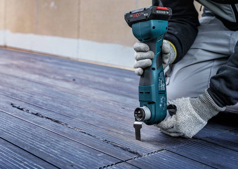 Eine Person repariert ein Holzdeck mit einem blauen Elektrowerkzeug. Sie trägt Handschuhe und arbeitet sorgfältig an den Fugen der Dielen.