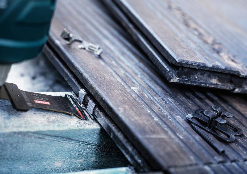 Nahaufnahme einer Säge, die durch dunkle Holzbretter schneidet, mit Metallklammern im Vordergrund. Szene von handwerklicher Arbeit.