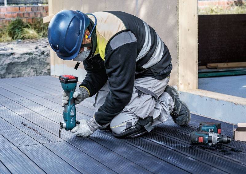 Arbeiter kniet auf Boden und benutzt einen Akkubohrer, um eine Holzplatte auf einem Deck zu befestigen. Trägt Schutzkleidung und Helm.