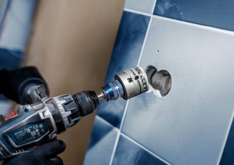 Handwerker bohrt mit einem leistungsstarken Werkzeug ein rundes Loch in eine glatte, blau-weiß geflieste Wand.