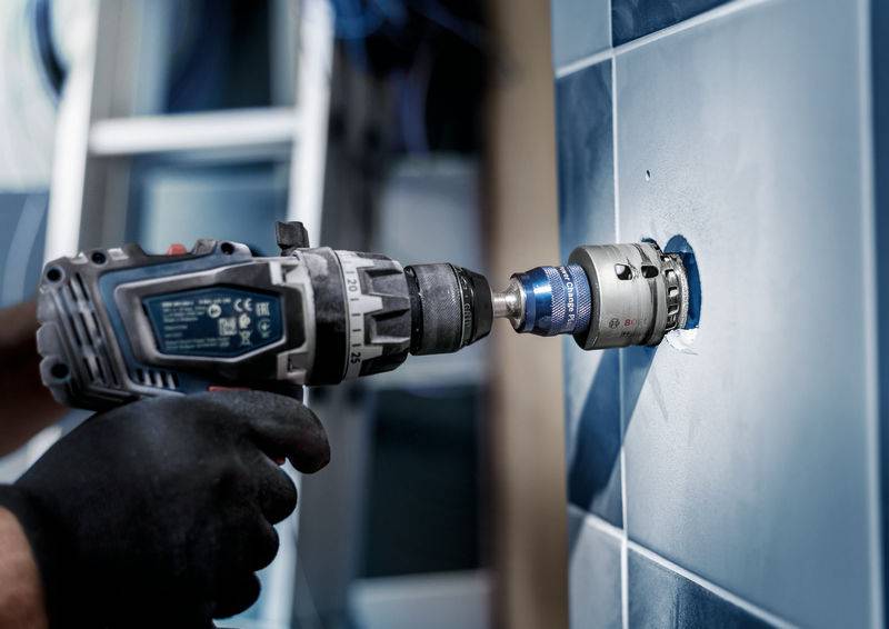 Eine Person bohrt mit einer elektrischen Bohrmaschine ein präzises Loch in eine geflieste Wand. Der Fokus liegt auf der Bohrmaschine.