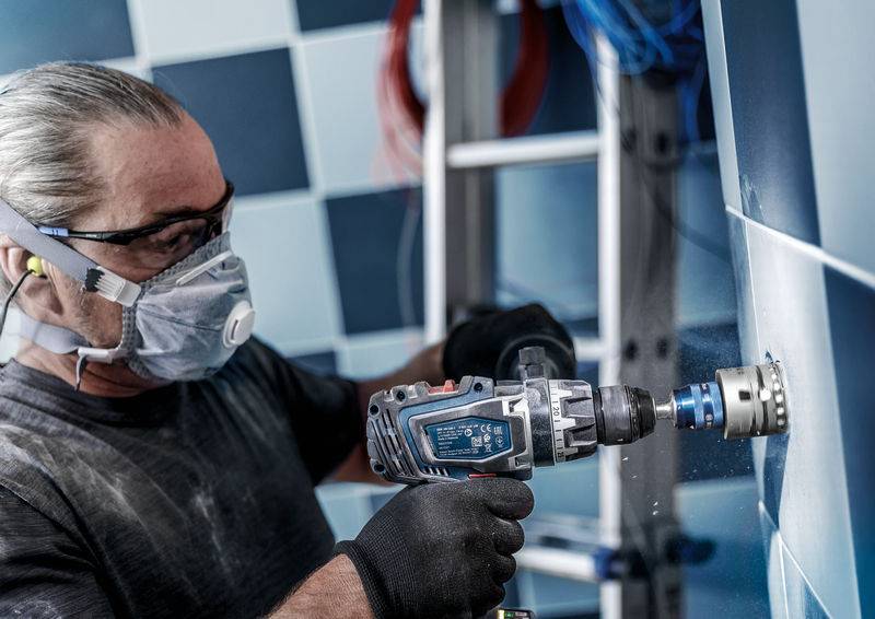 Ein Mann mit Schutzmaske und Brille bohrt mit einer Elektromaschine Löcher in eine geflieste Wand. Eine Leiter steht im Hintergrund.