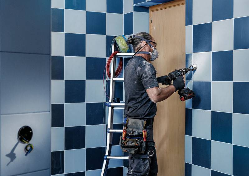 Eine Person mit Atemschutzmaske verwendet eine Bohrmaschine an einer gefliesten Wand. Im Hintergrund steht eine Leiter.