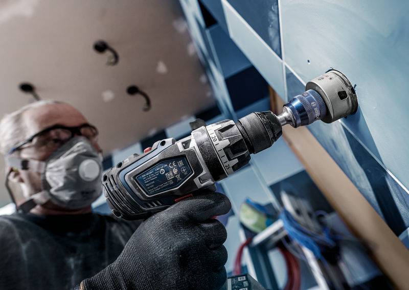 Eine Person mit Schutzbrille und Maske bohrt mit einer elektrischen Bohrmaschine ein großes Loch in eine geflieste Wand.
