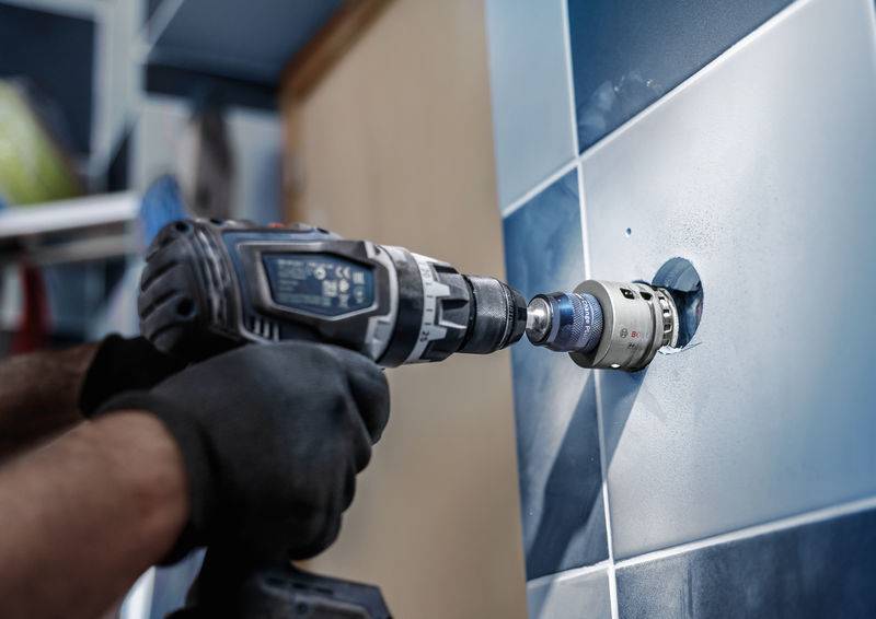 Eine Person bohrt mit einer Bohrmaschine ein Loch in eine blau geflieste Wand. Die Szene zeigt handwerkliche Arbeit.