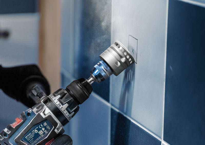 Eine Person bohrt mit einer Maschine ein kleines Loch in eine blaue Kachelwand. Die Fliese ist mit einem Bleistiftkreuz markiert.