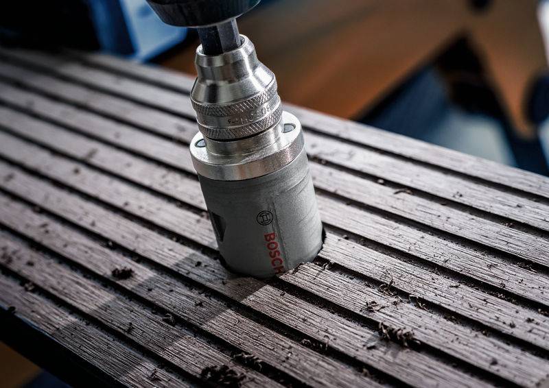 Ein Metallbohrer mit Bosch-Logo bohrt in ein geriffeltes Holzbrett. Holzspäne sind um die Bohrstelle verteilt.