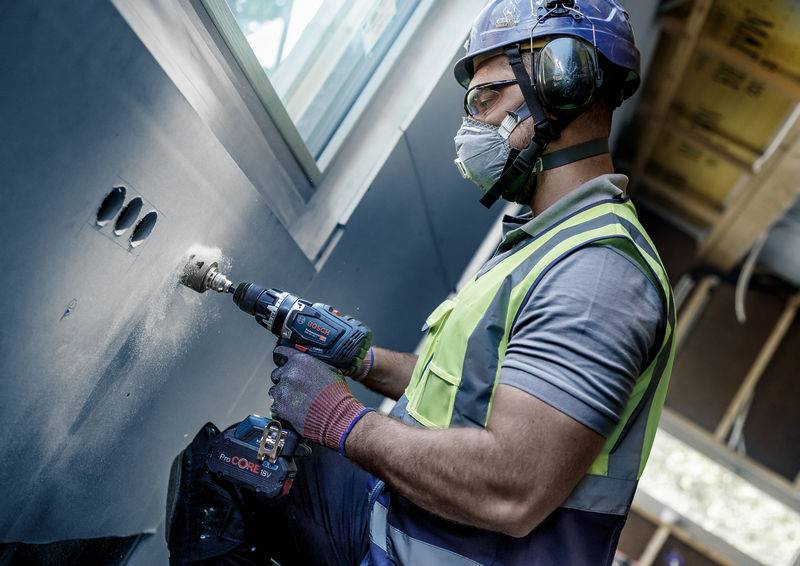 Ein Bauarbeiter mit Schutzkleidung bohrt präzise Löcher in eine Wand in einem teilweise fertiggestellten Raum.