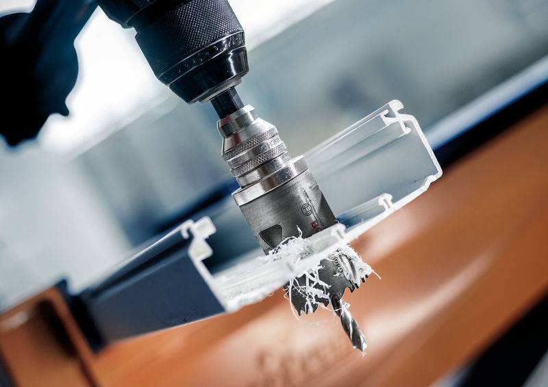 Bohrmaschine mit Lochsäge-Aufsatz schneidet durch ein Metallprofil. Späne fliegen in die Luft. Fokus auf den Bohrvorgang.