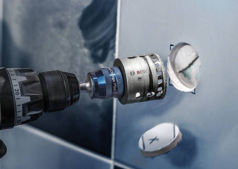 Ein Bohrer mit Aufsatz bohrt Löcher in blaue Fliesen. Ein bereits gebohrtes Loch daneben.