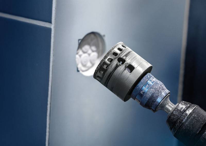 Ein elektrischer Bohrer mit Lochsägeaufsatz wird auf eine geflieste Wand gerichtet, neben einem gebohrten Loch mit sichtbarer Steckdose.