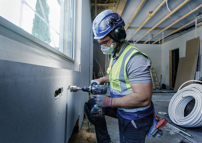 Ein Bauarbeiter mit Helm und Schutzbrille bohrt ein Loch in eine Wand in einem halbfertigen Raum, umgeben von Baumaterialien.