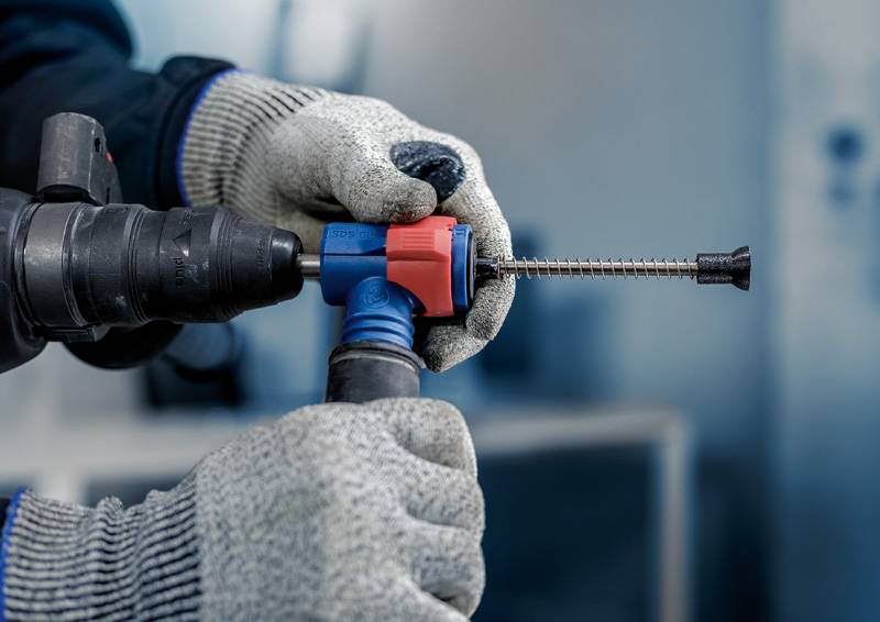 Person mit Handschuhen hält eine Bohrmaschine, die einen langen Schraubanker in eine Wand setzt. Fokus auf Werkzeug und Schraube.