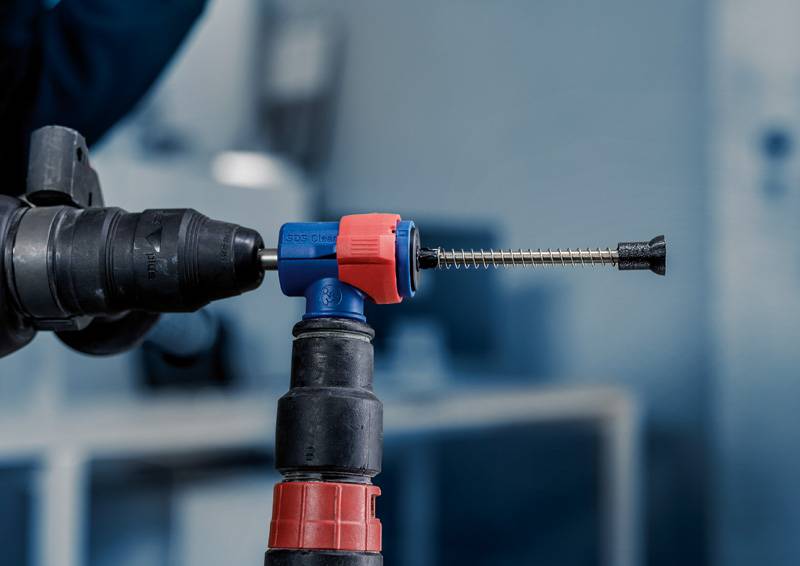 Ein Bohrer mit blau-rotem Aufsatz und schwarzem Bohrer im Fokus, vor unscharfem Hintergrund in einer Werkstattumgebung.