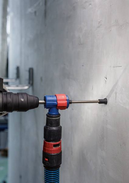 Eine Bohrmaschine wird benutzt, um ein Loch in eine graue Wand zu bohren. Ein Staubabsauger ist an der Bohrstelle montiert.
