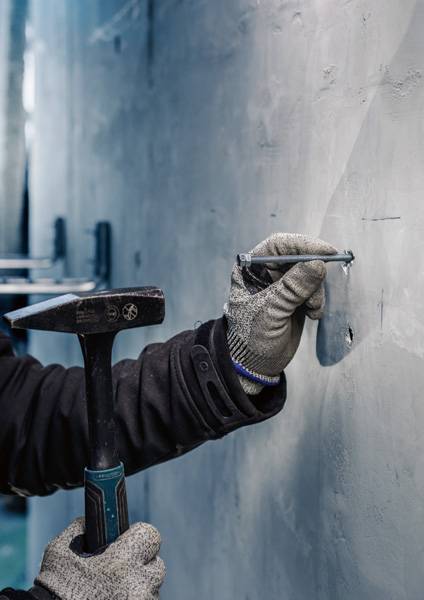 Eine Person mit Handschuhen hämmert einen Nagel in eine graue Betonwand. Die Umgebung wirkt industriell.