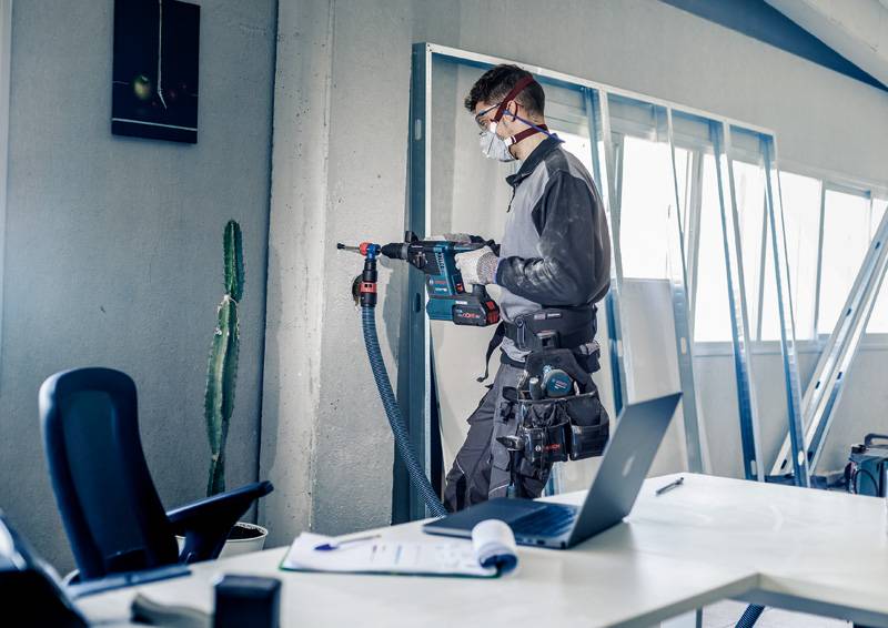 Ein Handwerker mit Schutzbrille und Maske bohrt eine Wand in einem Büro. Auf dem Tisch liegt ein Laptop, ein Ordner und ein Stift.