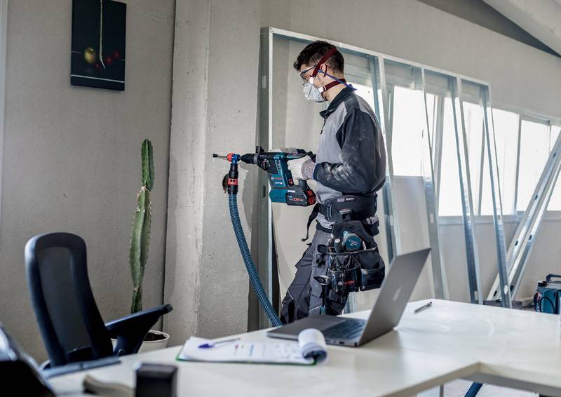 Ein Handwerker mit Schutzbrille und Maske bohrt mit einer elektrischen Bohrmaschine in die Wand in einem Büro mit Tisch und Laptop.