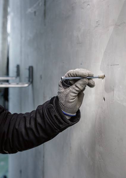 Eine behandschuhte Hand hält einen Bleistift und markiert eine Wand aus grauem Beton.