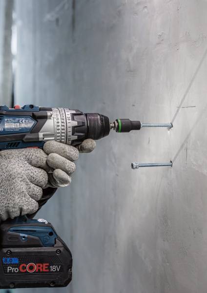 Person in Arbeitshandschuhen verwendet eine Bohrmaschine, um Schrauben in eine graue Wand zu bohren. Elektrisches Bohrwerkzeug in Nahaufnahme.