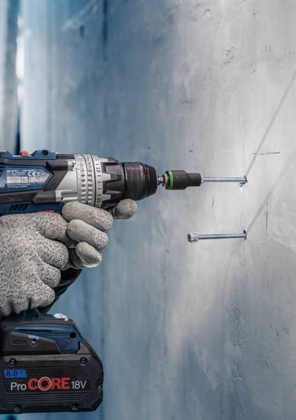 Eine Nahaufnahme einer Person, die mit einer blauen Akkubohrmaschine von Bosch in eine Betonwand bohrt, während zwei Schrauben befestigt sind.