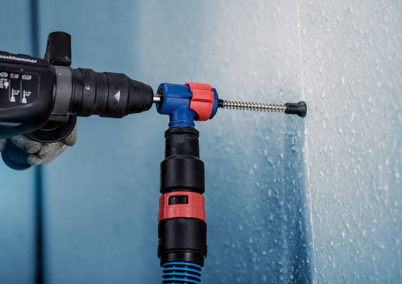 Eine Person bohrt mit einem Bohrhammer in eine blaue Wand. Der Bohrer hat eine Staubabsaugung, die den Bohrstaub aufnimmt.