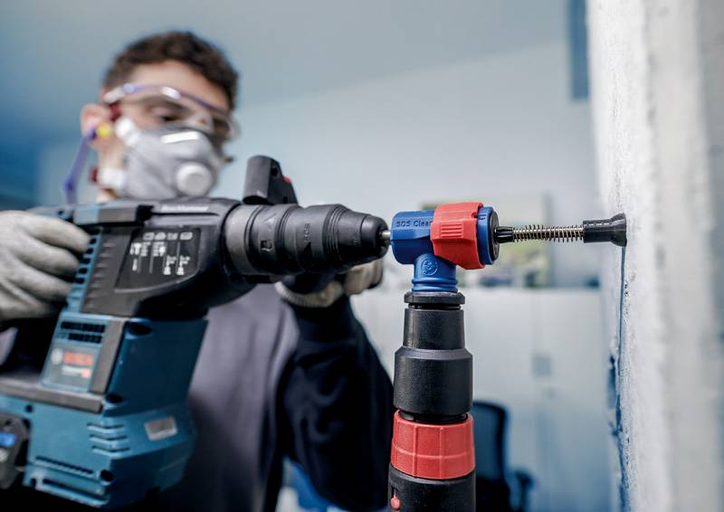 Eine Person trägt Schutzbrille und Atemmaske, während sie mit einer Bohrmaschine an einer Wand arbeitet. Sie hält das Werkzeug mit beiden Händen.